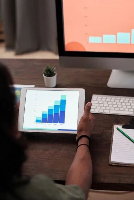 Hands using smartphone beside laptop with stock charts, showcasing digital trading.